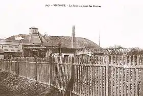 Carte postale ancienne en noir et blanc de la fosse n° 4 des mines de Vicoigne à Raismes et montrant en arrière-plan la fosse avec son bâtiment d'extraction et son chevalement recouvert de bois, et son terril plat. Une clôture est visible au premier plan.
