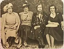 quatre dames assises sur un banc, Victoria-Eugénie en tenue claire portant un chapeau à plumes, Henriette en tenue claire coiffée d'un petit chapeau, Marie-José vêtue d'une tenue sombre à motifs clairs et Lilian, souriante en tenue sombre et tenant un livre sur ses genoux