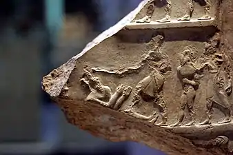 Photographie d'un fragment de sculpture en bas-relief, représentant quatre personnages dont deux sont habillés et les deux autres pas. Aux endroits où elle est intacte, la sculpture présente des traits détaillés précis au niveau des vêtements et de l'anatomie.
