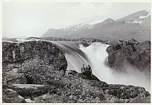 Photo en noir et blanc avec deux hommes assis devant une grosse chute d'eau, au milieu des montagnes.
