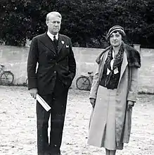 Un homme en costume à l'air sévère et une femme souriante vêtue d'une robe longue, d'une redingote et d'un chapeau.