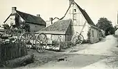 Carte postale en noir et blanc de bâtiments agricoles, au premier plan figurent une charrette à bras et des roues de charrette