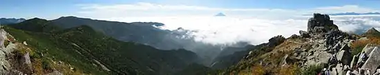 Photo couleur montrant le sommet conique d'un volcan, émergeant d'une couche de nuages blancs au loin, à l'horizon, depuis la cime d'une montagne.