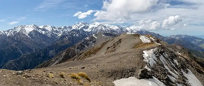 Panorama de sommets enneigées de montagnes depuis une haute colline de terres nues.