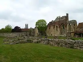 Des bâtiments en ruine (au premier plan ne restent que les fondations, des murs se dressent au second plan). On voit la tour d'une cathédrale à l'horizon.