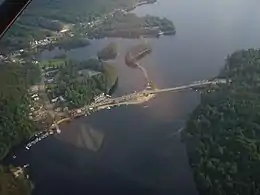 A large lake is surrounded by land partially or wholly covered in forests. A road crosses the lake via bridge and intersects another that crosses a small bay by way of a narrow strip of land. At lower left is a community composed mostly of houses. The highway carried by the bridge across the lake serves the community and intersects another highway at upper left.