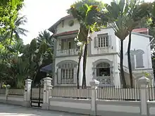 Façade d'une maison blanche à deux étages, entourée de palmiers.