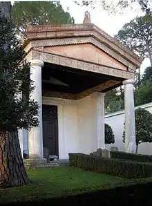 Photographie de la reconstitution, dans un parc boisé, d'un petit temple étrusque avec un fronton porté par deux colonnes et une architrave avec des bas-reliefs.