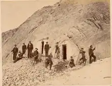 Photographie montrant un groupe d'hommes posant devant un abri creusé dans la montagne.
