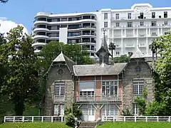 Photographie en couleurs d'une villa dont la toiture porte un clocheton central ; immeuble moderne en arrière-plan.