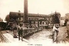 Vue d'une usine et de sa cheminée au bord d'un cours d'eau sur une carte postale des années 1900.