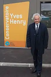 Photo d'un homme posant devant un bâtiment ; panneau portant son nom « Yves Henry ».