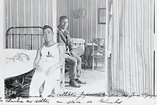 Photo en noir et blanc de deux hommes dans une chambre. Du texte, illisible, est écrit à la main en bas de la photo.