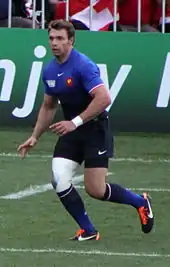 De trois-quarts, un homme au maillot bleu, le genou droit bandé, court sur un terrain de rugby.