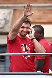 Photographie couleur d’un homme, de face, vêtu d’un t-shirt rouge, au téléphone, faisant signe avec le bras droit tendu. En arrière-plan, un autre homme vêtu du même t-shirt.