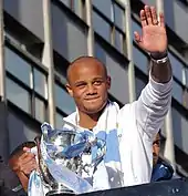 Vincent Kompany, de face, lors de la célébration de fin de saison 2013-2014. Il tient le trophée de la Coupe de la ligue d'Angleterre dans la main droite et fait un signe à la foule de la main gauche.