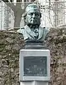 An outdoor bust of a bearded man wearing a large patterned cap and a dicky bow tie. A plaque beneath reads: Vincent Wallace composer. Born Waterford 1812, died Pyrennes 1865. "In happy moments day by day, The sands of life may pass. In swift but tranquil tide away, From times inferring glass."