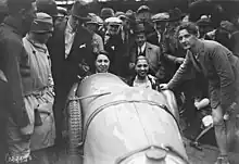 Photographie en noir et blanc d'une voiture de course métallique entourée par une foule, avec deux femmes à son bord, la conductrice fume.