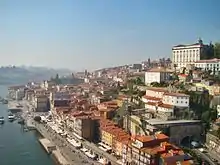 Une ville côtière très ensoleillée sur la droite, construite à flanc de colline. Les nombreuses maisons blanches, jaunes ou rouges ont toutes des toits de tuiles oranges. L'eau d'un estuaire et quelques petits bateaux sont visibles au pied de la colline sur la gauche