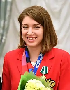 Portrait d'une femme, souriante, en tenue, décorée d'une médaille, portant un bouquet de fleurs devant elle.