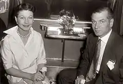Photo en noir et blanc d'un homme et d'une femme. La femme est à gauche, les mains sur les genoux, souriante. L'homme est en costume cravate à droite, l'air sérieux. Ils ne se touchent pas.