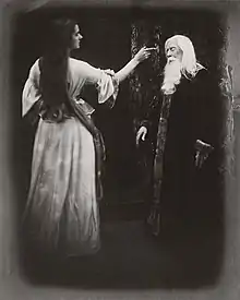 Une jeune femme en robe, à gauche, tend un doigt vers le front d'un vieil homme barbu, à droite.