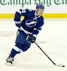 Photographie couleur d’un hockeyeur de pied, de face, penché sur sa gauche, vêtu à dominante de bleu, regardant l’objectif de côté