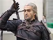 Photographie couleur d'un homme à la chevelure argentée vêtu de lourds vêtements, protections et gantelets de cuir, l'épée sur l'épaule et un médaillon en forme de loup autour du cou.