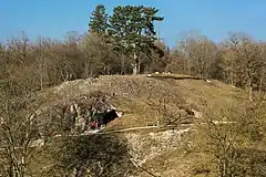 Photographie en couleurs d'une colline percée de deux ouvertures, recouverte d'un tapis herbeux, d'arbres et de roches affleurantes, un étroit sentier aménagé d'un banc visible à mi-hauteur.