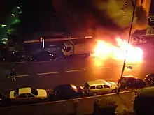 De nuit, une voiture en feu dans une rue.