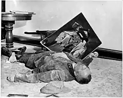Photo noir et blanc prise le 19 avril 1945, à l’intérieur d’un bureau de l’hôtel de ville de Leipzig. Sur le sol, un homme mort, en uniforme, est allongé de tout son long, sa tête vers la droite au premier plan. Un mur blanc forme l’arrière-plan de la photo. Au second plan, entre une chaise renversée le long du mur et le flanc droit du cadavre, tient en équilibre un portrait d’Adolf Hitler dont le visage a été lacéré.