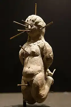 Statuette en terre d'une femme nue, poignets et chevilles liés dans le dos, percée d'épingles dans le crâne, les oreilles, les yeux, la bouche, le sternum, le sexe et les pieds