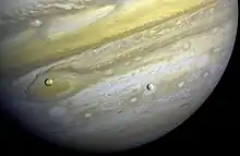 Le fond de l'image est occupé par Jupiter. Deux sphères sont devant la planète, une orangée (Io) vers la gauche et une grisâtre (Europe) vers le centre.