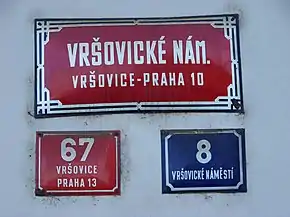 Trois plaques de rues : l'une en haut, très grande, horizontale et rouge indique le nom de la rue, ainsi que celui du quartier et de l'arrondissement ; en dessous, une petite plaque rouge mentionne un numéro, le nom du quartier et d'un autre arrondissement ; à sa droite, une autre petite plaque bleue indique un autre numéro et le nom de la rue.