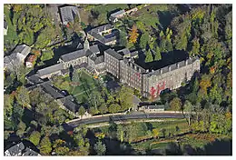 Photographie aérienne d'un paysage arboré montrant un grand bâtiment à plusieurs ailes, bodé au premier plan d'un ruisseau et d'une route.