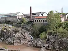 photo couleur montrant un chaos sur une rivière. Sur l'autre rive, plusieurs longs bâtiments industriels et deux hautes cheminées constituent une usine.