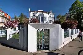 Vue du portail piétonnier et du muret d'enceinte d'une villa, aux formes anguleuses, le tout dans des tons blancs