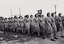 Un groupe de femmes en uniforme défile. Elles portent un petit étendard avec le symbole du corps des ingénieurs ; un château surmonté de la lettre D.
