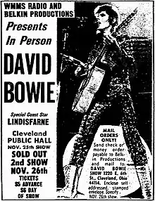 Photographie en noir et blanc d'une affiche annonçant un concert de David Bowie à Cleveland. À droite le chanteur en pied, vêtu d'un justaucorps et de bottes, guitare à la main.