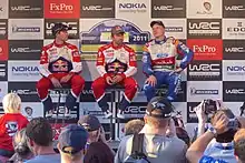 Sébastien Loeb avec un micro, entouré de Sébastien Ogier et Jari-Matti Latvala, tous trois assis sur un tabouret et faisant face à des spectateurs.