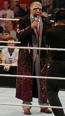 Un catcheur en costume et micro à la main parle au milieu d'un ring.