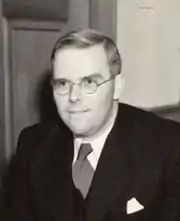 photo noir et blanc d'un jeune homme blond, en costume sombre, avec cravate claire et lunettes rondes