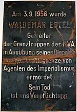 Plaque de métal endommagée indiquant « Am 3.9.1956 wurde WALDEMAR ESTEL Getreiter der Grenztruppen der NVA in Ausübung seine Dienstes an der Staatsgrenze von Agenten des Imperialismus ermordet. Sein Tod ist uns Verpflichtung. »