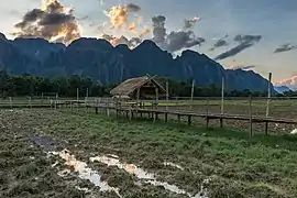 Passerelle en bois sur pilotis menant vers une hutte au toit de paille au milieu des rizières avec réflexion dans l'eau des nuages colorés au coucher du Soleil, et des montagnes karstiques dans la campagne de Vang Vieng, durant la mousson.