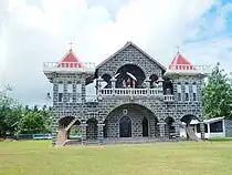 Bâtiment en pierres noires avec des jointures blanches, sur deux niveaux, flanqué de deux petites tours au toit rouge. Au premier plan, une pelouse.