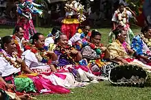 Photographie d'un groupe de femmes assises dans l'herbe, en tenue traditionnelles (jupes aux couleurs vives, colliers de fleurs). Devant elles est disposé un panier tressé contenant des billets, et certaines d'entre elles ont des billets épinglés dans les cheveux.