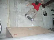 Un skater effectue un wallride contre un mur en béton. Il a pris son élan sur une toute petite rampe placée au bas du mur et est en train de rouler avec sa planche contre le mur.