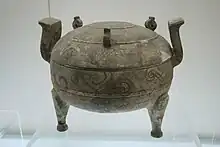 Photographie montrant une poterie de forme sphérique, supportée par 3 pieds (2 visibles uniquement) et ornée de deux poignées diamétralement opposées sur le haut de l'objet.