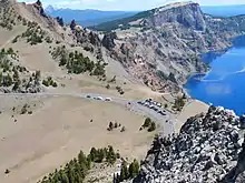 Vue plongeante depuis des rochers sur une route et un parking dominant le lac cerné par des reliefs déchiquetés.