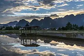 Réflexion dans l'eau d'une hutte en bois avec un ciel coloré de nuages oranges, gris et blancs, dans l'eau d'une rizière, avec des montagnes karstiques au coucher du soleil pendant la mousson, dans la compagne de Vang Vieng.